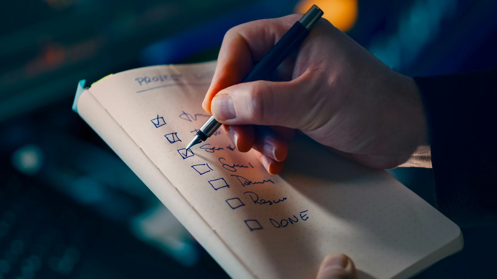A hand with a fountain pen ticks boxes on a handwritten checklist in a dotted notebook, against a dark, softly lit background