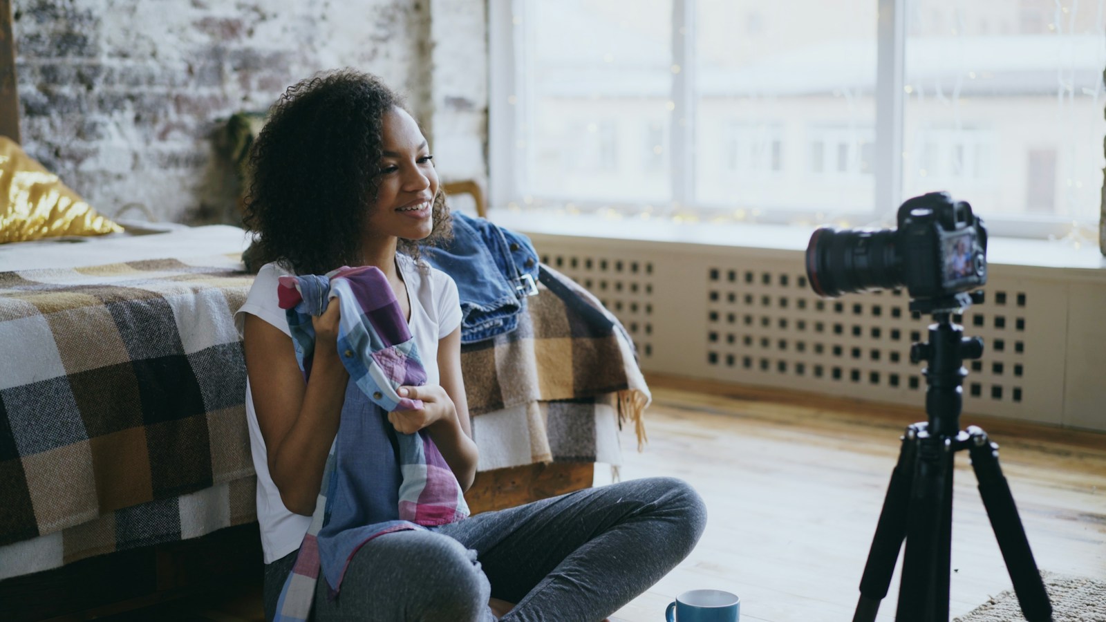 A smiling woman sitting on a loft floor holds up a plaid shirt to a camera on a tripod while filming a clothing video