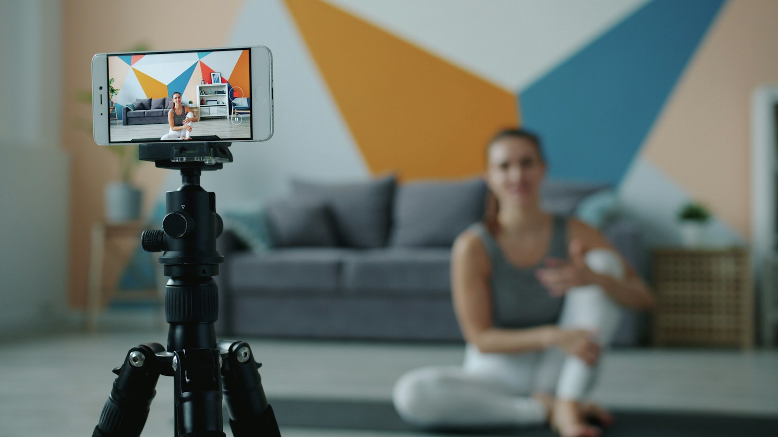A smartphone on a tripod records a woman sitting on a yoga mat as she talks to the camera, her image framed on the phone’s screen