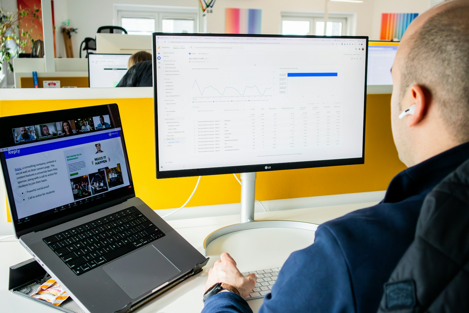 Over-the-shoulder view of a man at a large monitor showing an analytics report, with a laptop open beside him, comparing results across two screens