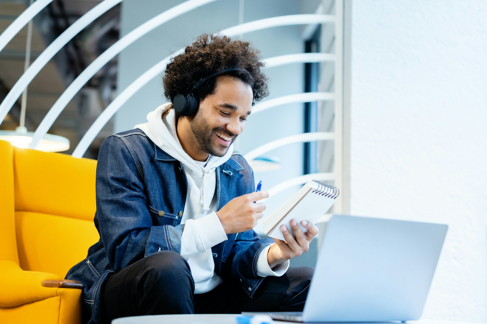 A man in headphones smiles while writing notes in a spiral notepad, watching a video on an open laptop
